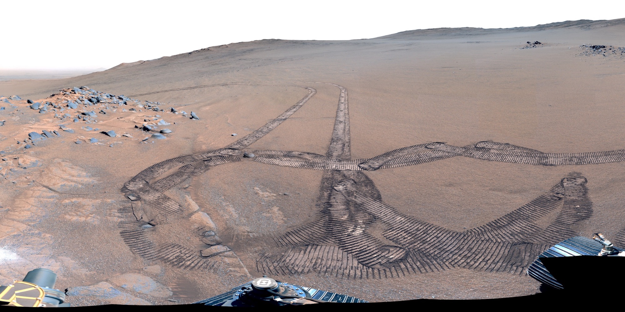 Rover wheel tracks looping across reddish Martian sand, with rocky outcrops and a low hill under a pale sky